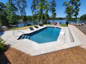 a custom fiberglass pool with an asymmetrical design, installed by Kirk's Pool and Hearth. There are tall trees and a lake in the background